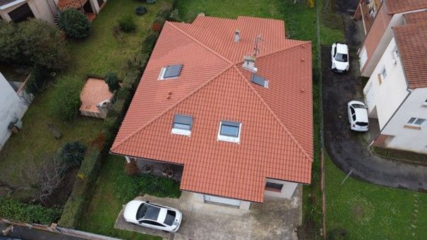 Vue aérienne d'une maison au toit de tuiles rouges, avec quatre puits de lumière et des voitures garées dans l'allée et dans la rue latérale.