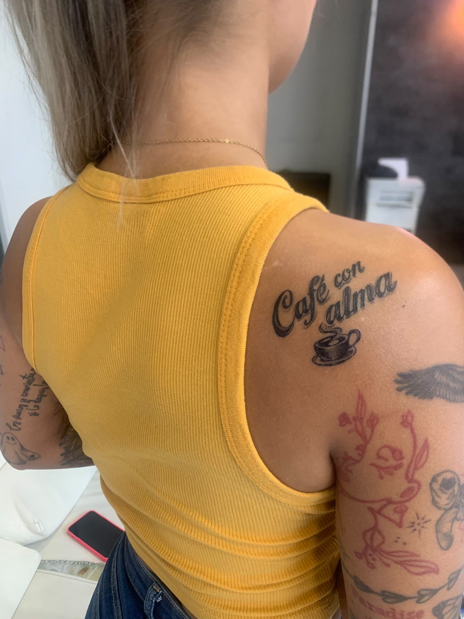 Vista trasera de una persona con una camiseta amarilla sin mangas y tatuajes visibles en ambos brazos y hombros.