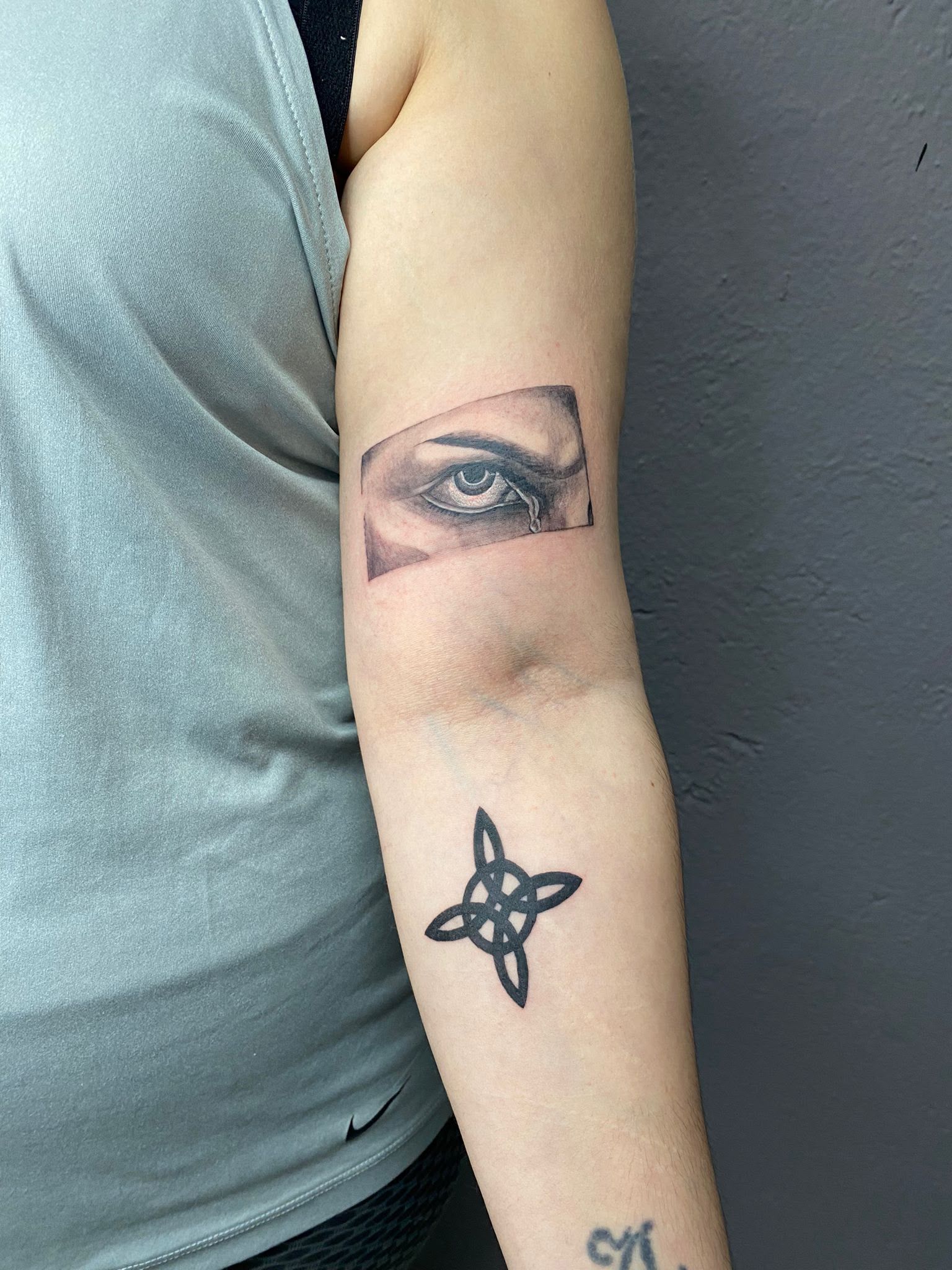 Brazo con tatuaje de ojo negro y pequeña estrella, sobre una camiseta y una pared grises