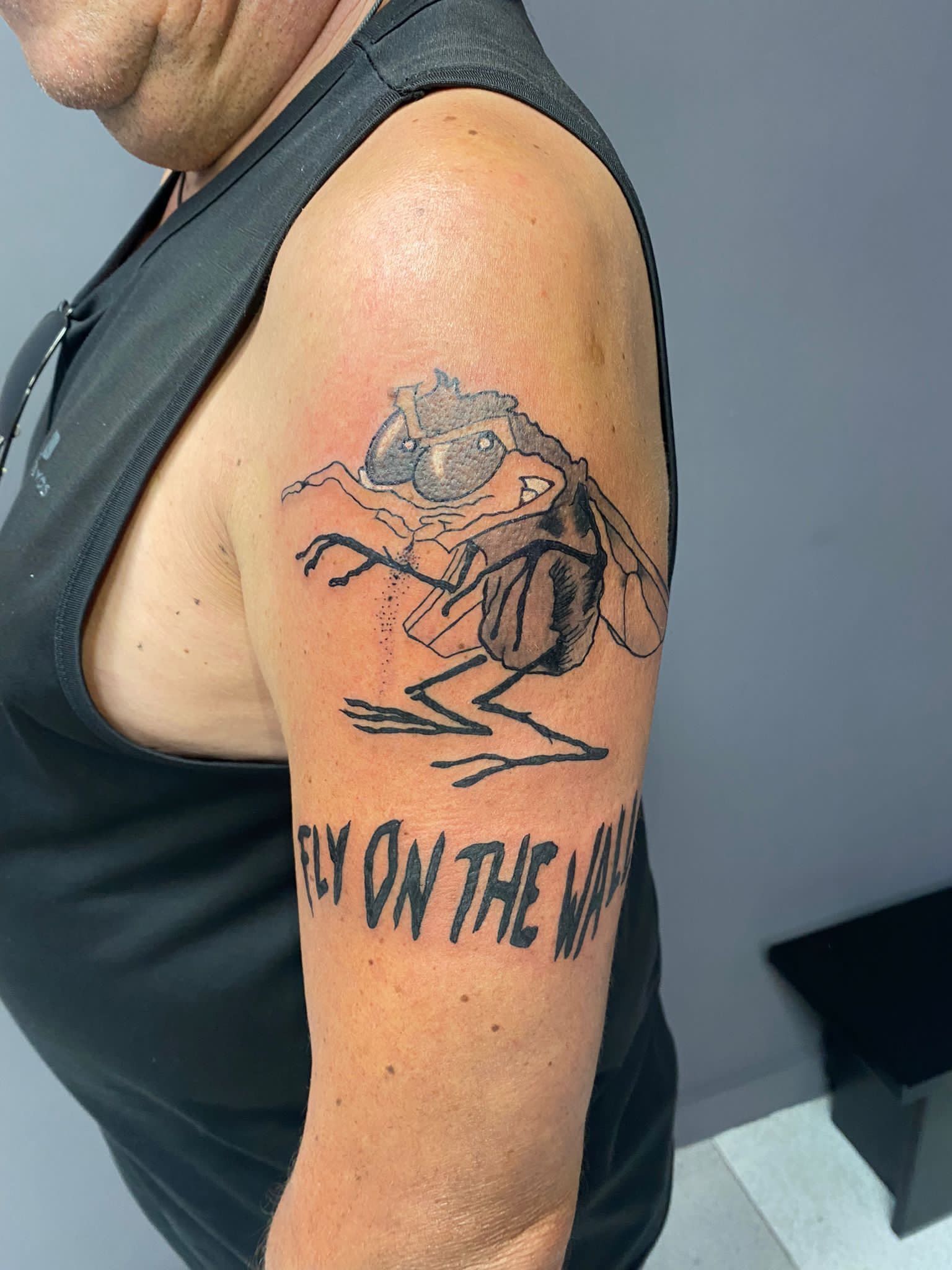 Primer plano de la parte superior del brazo con un tatuaje negro y texto parcialmente visible en una camiseta sin mangas.