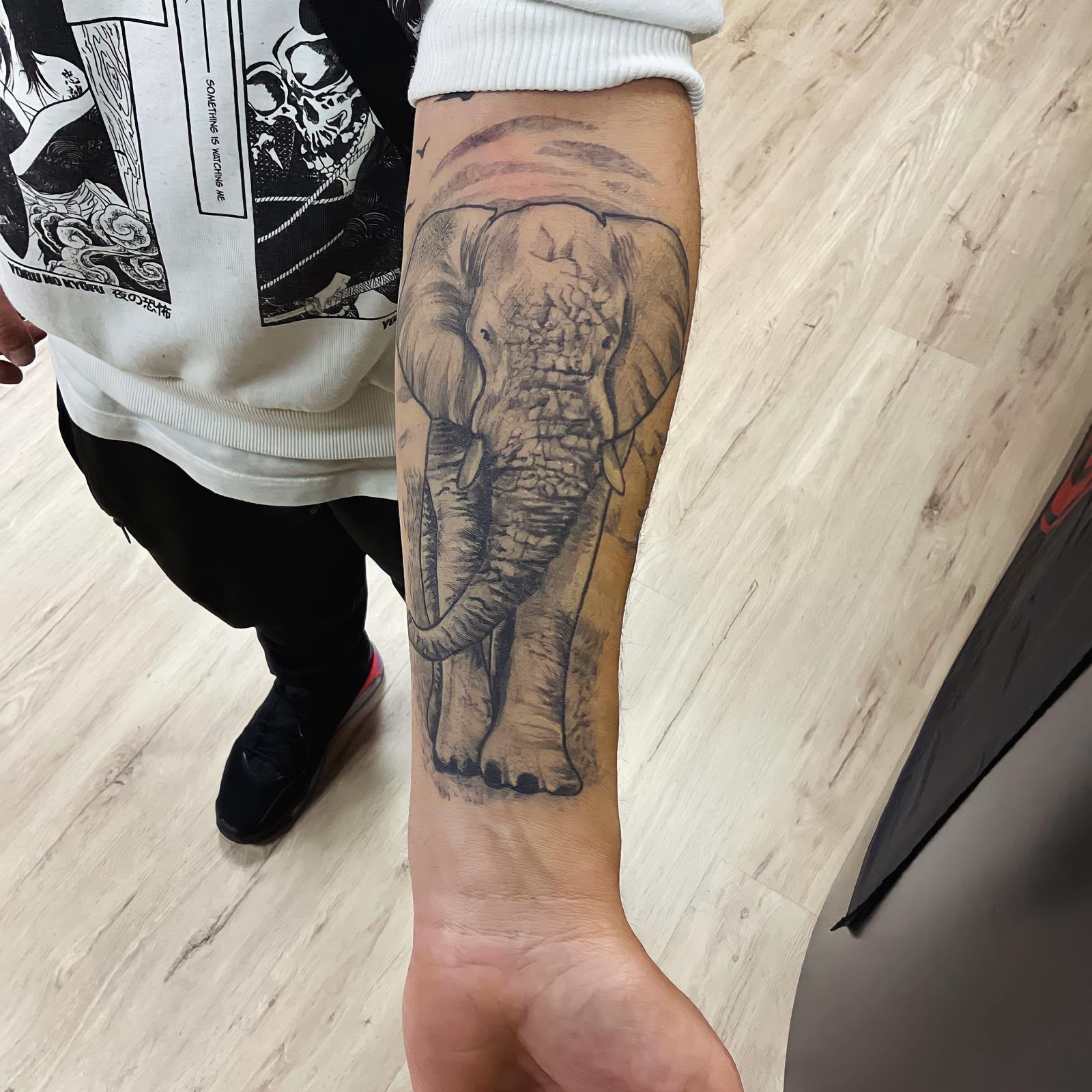 Brazo con un tatuaje de elefante en una habitación luminosa, con una sudadera blanca y pantalones negros.