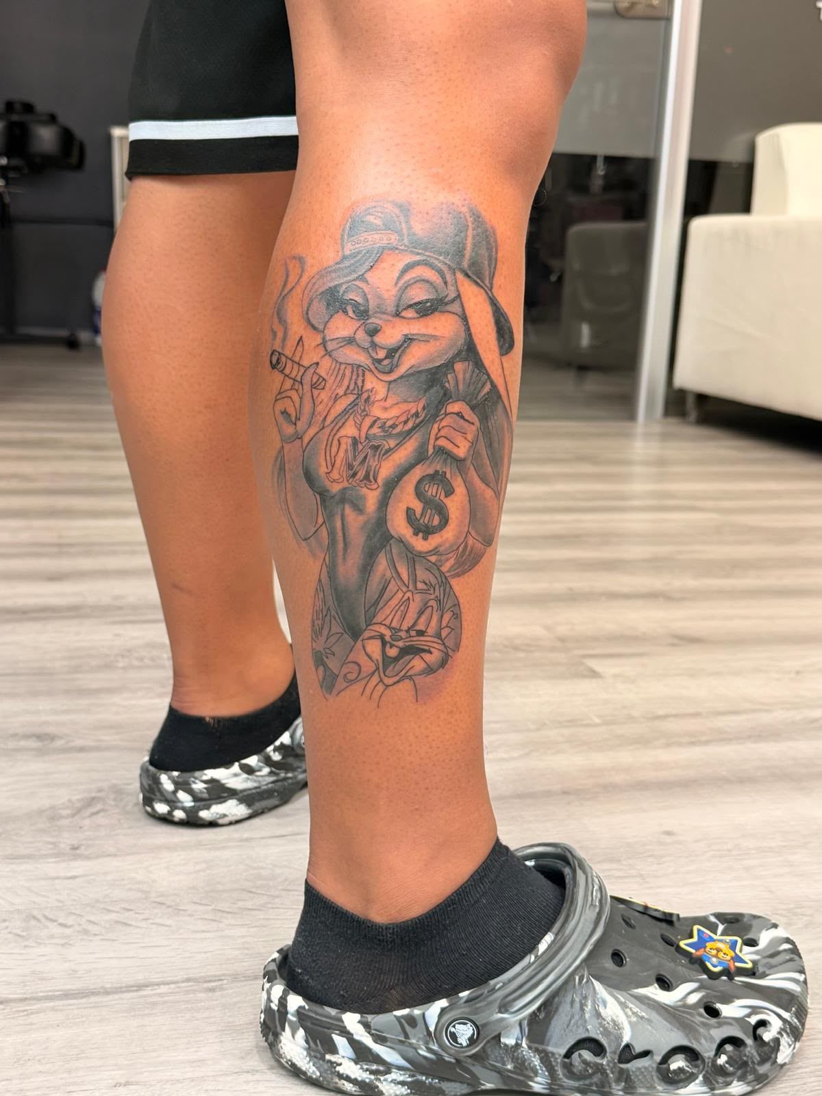 Pierna con un tatuaje en blanco y negro de una mujer y una rosa, con calcetines negros y Crocs de camuflaje en interiores