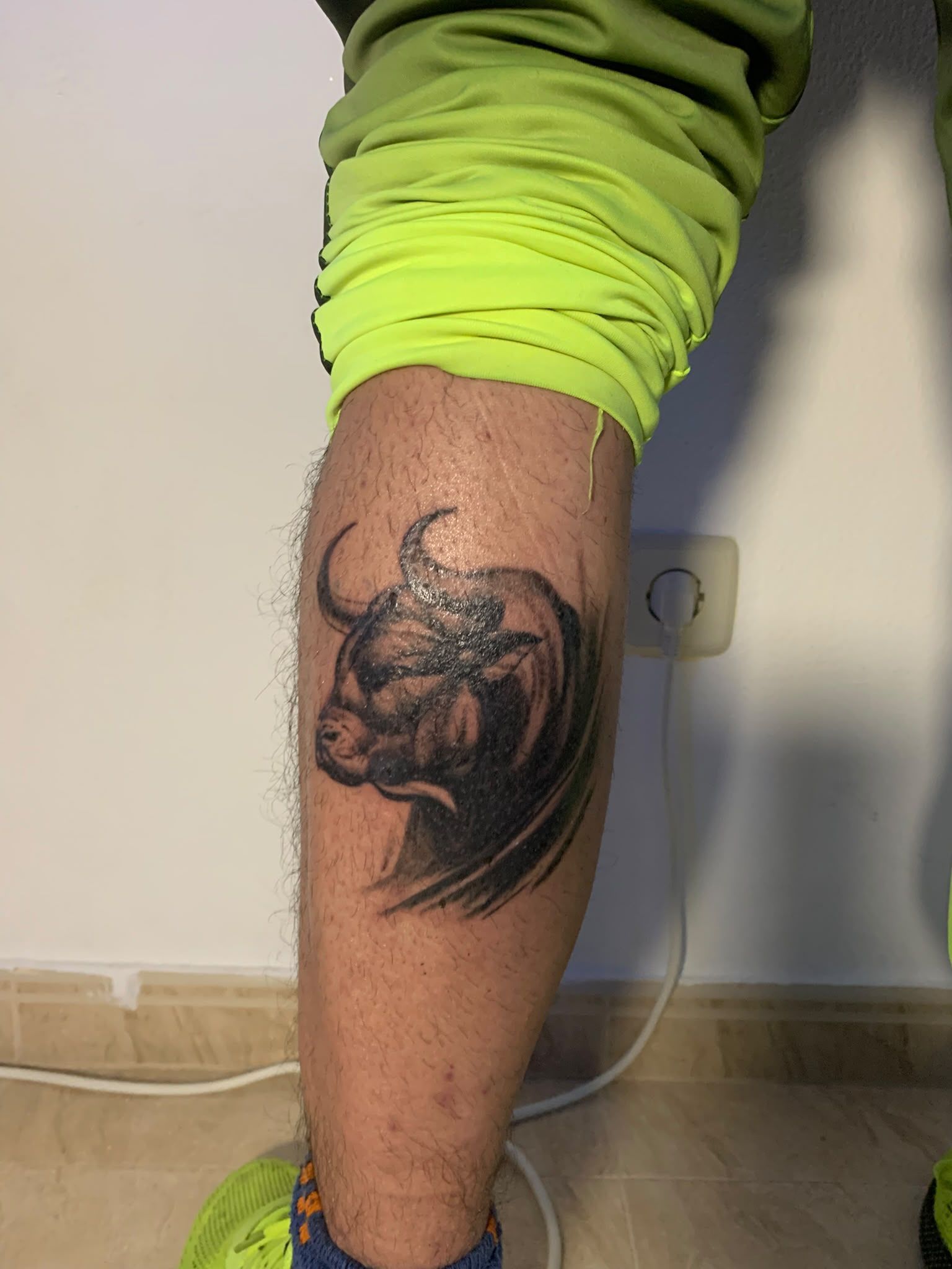 Parte inferior de la pierna, con un tatuaje de un toro negro cerca de la pantorrilla, en un interior sobre un suelo de baldosas.