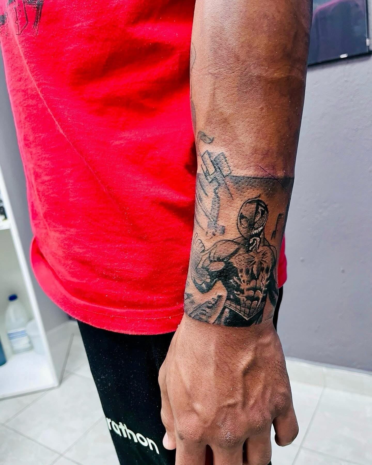 Brazo con un tatuaje de manga negra junto a una camiseta roja y unos pantalones negros, frente a una pared gris