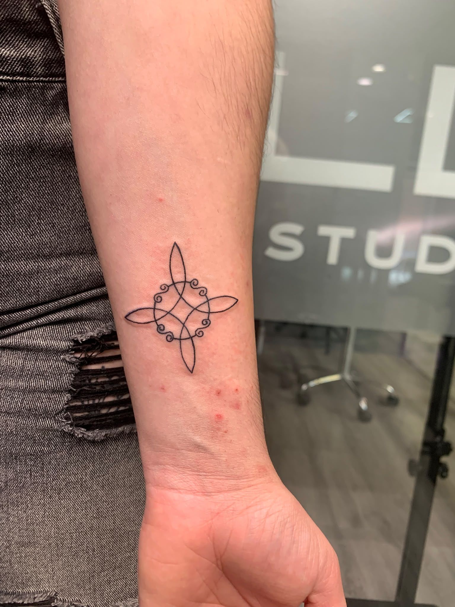 Antebrazo con un pequeño tatuaje floral negro cerca de la muñeca, delante de una puerta de cristal.