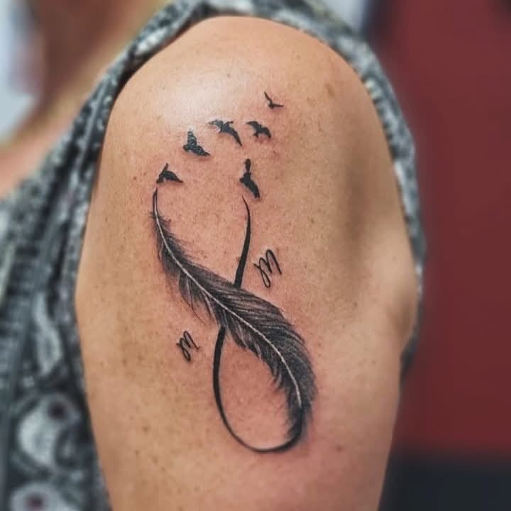Tatuaje de una pluma negra en forma de infinito en el hombro, con pequeños pájaros volando.
