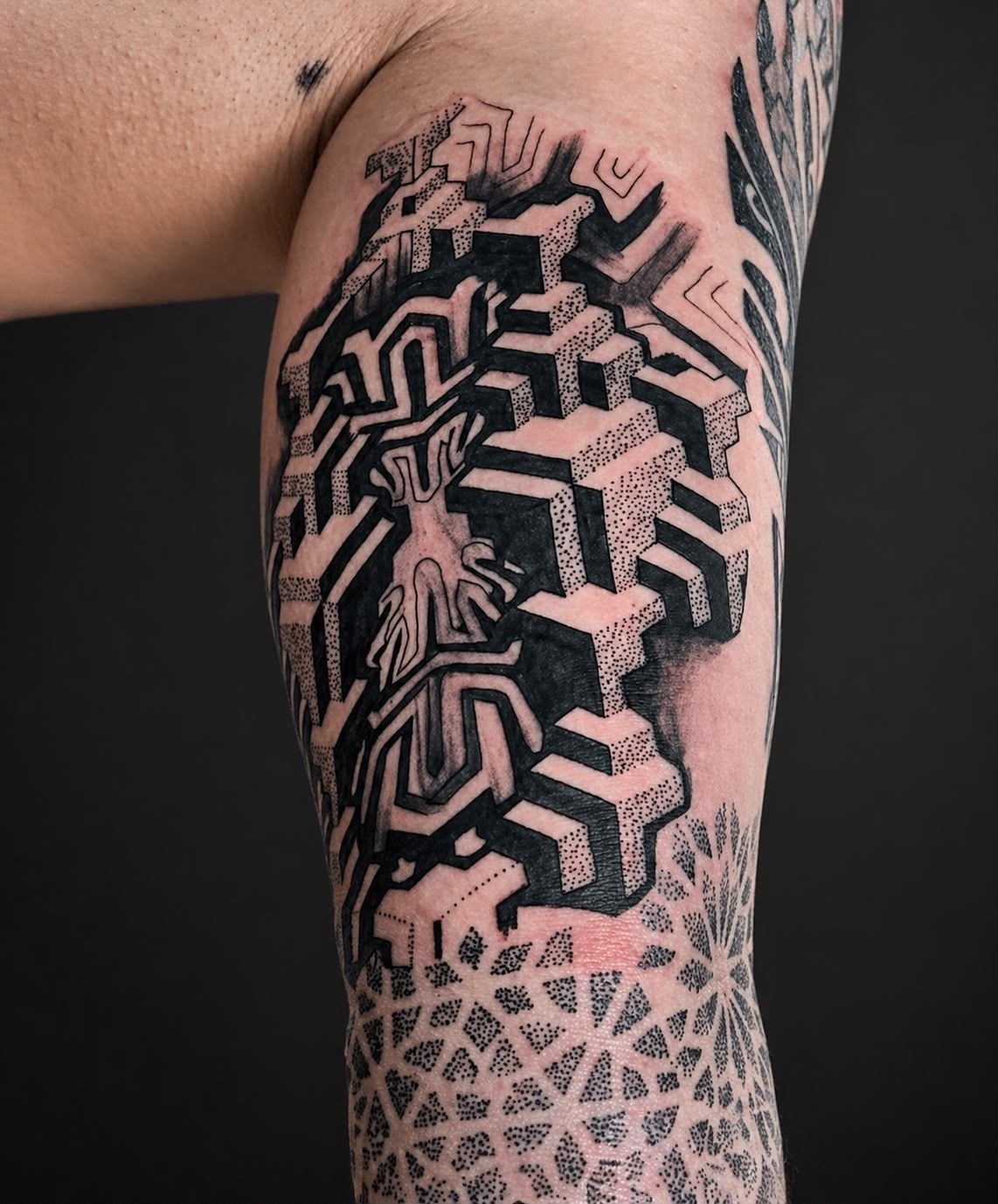 Primer plano de un tatuaje geométrico negro en la parte superior del brazo de una persona, con la piel de debajo decorada con patrones