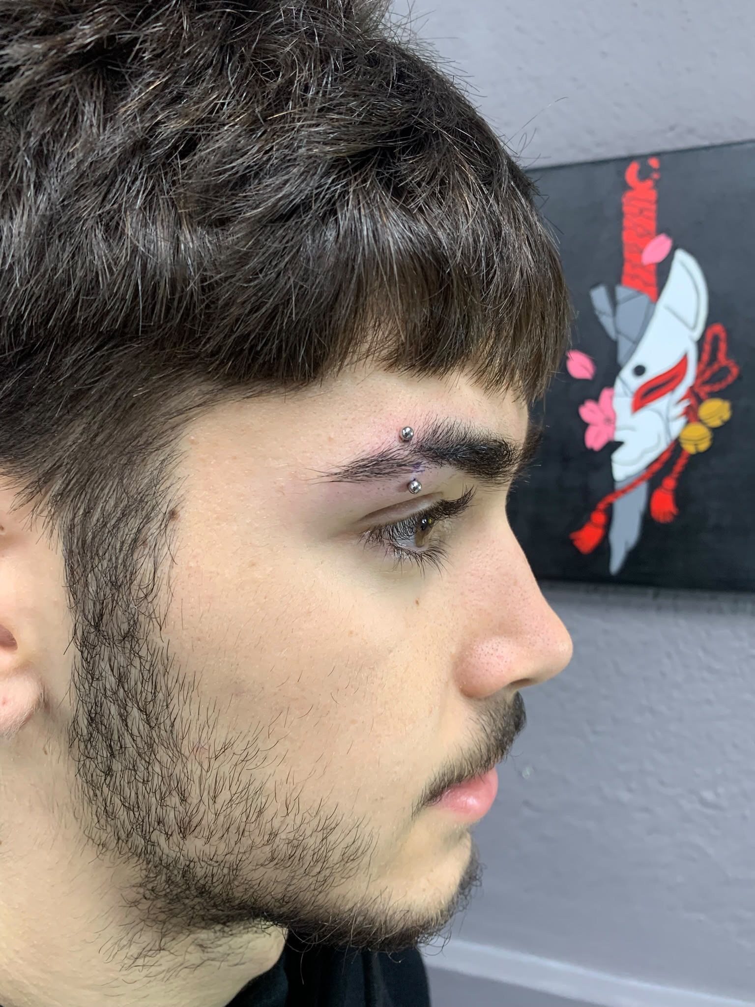 Perfil de una persona de cabello oscuro y barba clara, frente a una pared gris con una pequeña obra de arte colorida colgada en la pared.