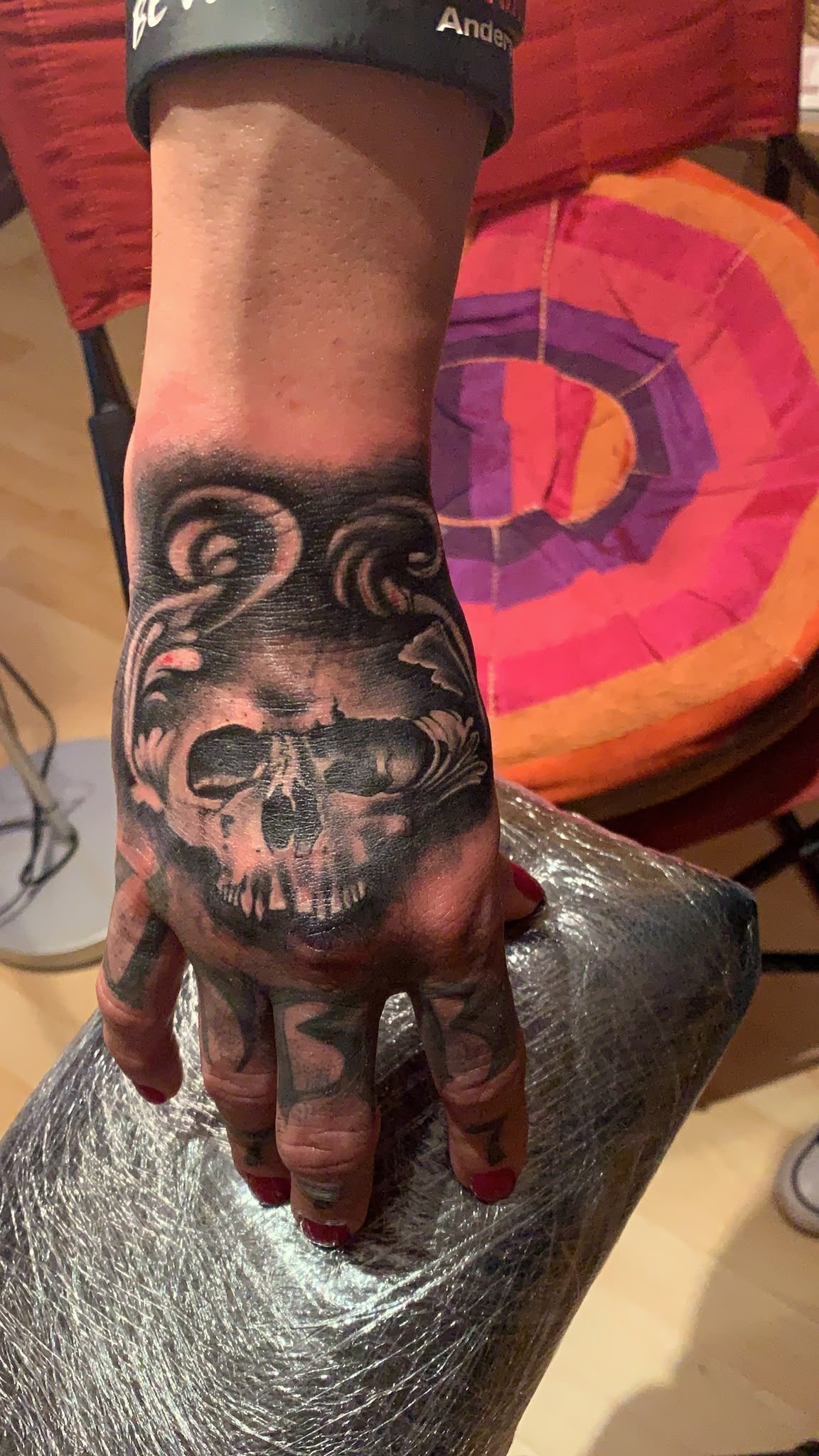 Mano con un tatuaje de una calavera negra apoyada en una silla plateada, con un cojín circular de colores al fondo