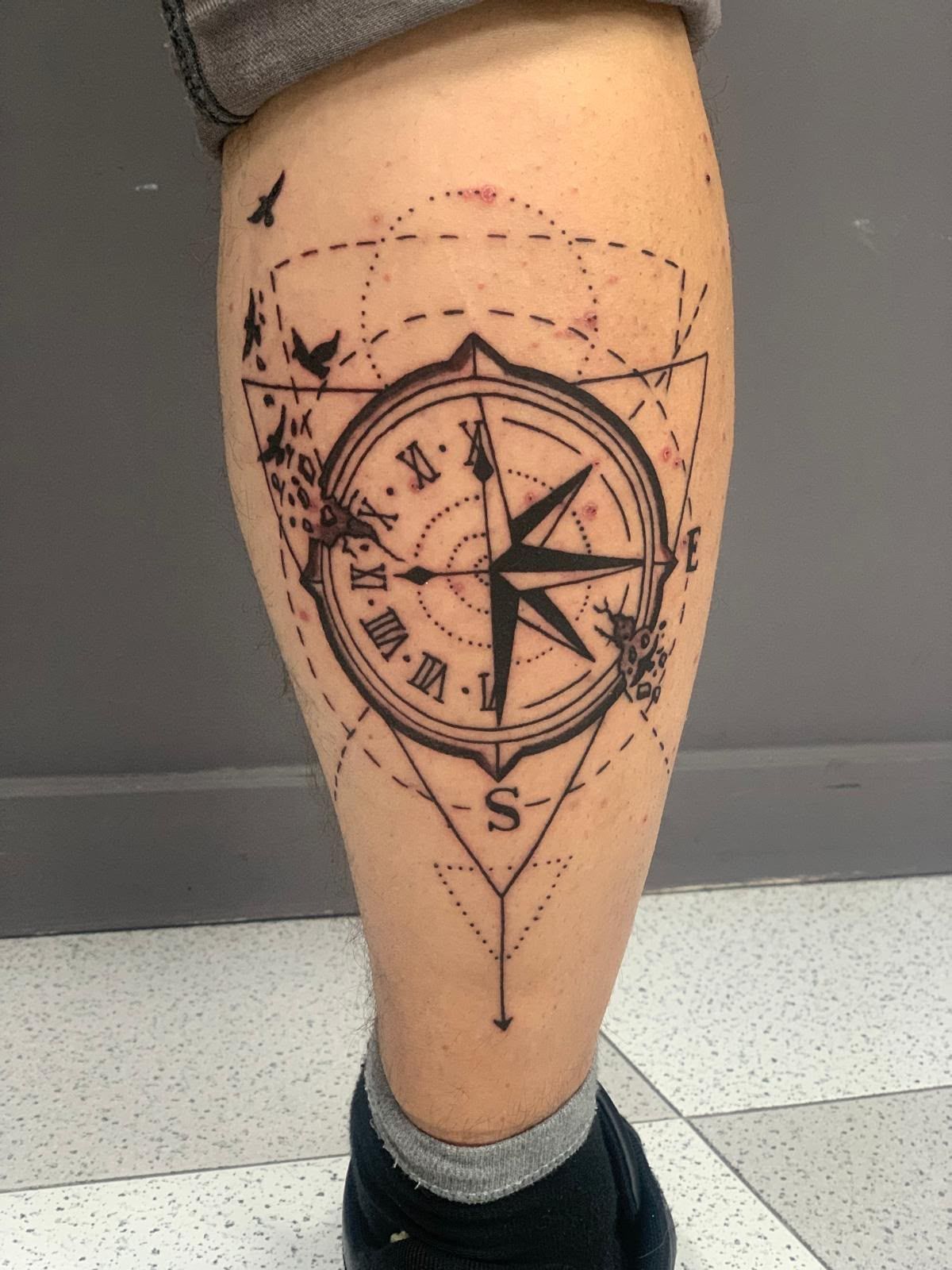 Tatuaje de brújula en la parte inferior de la pierna, en tinta negra, con diseño náutico sobre un fondo claro