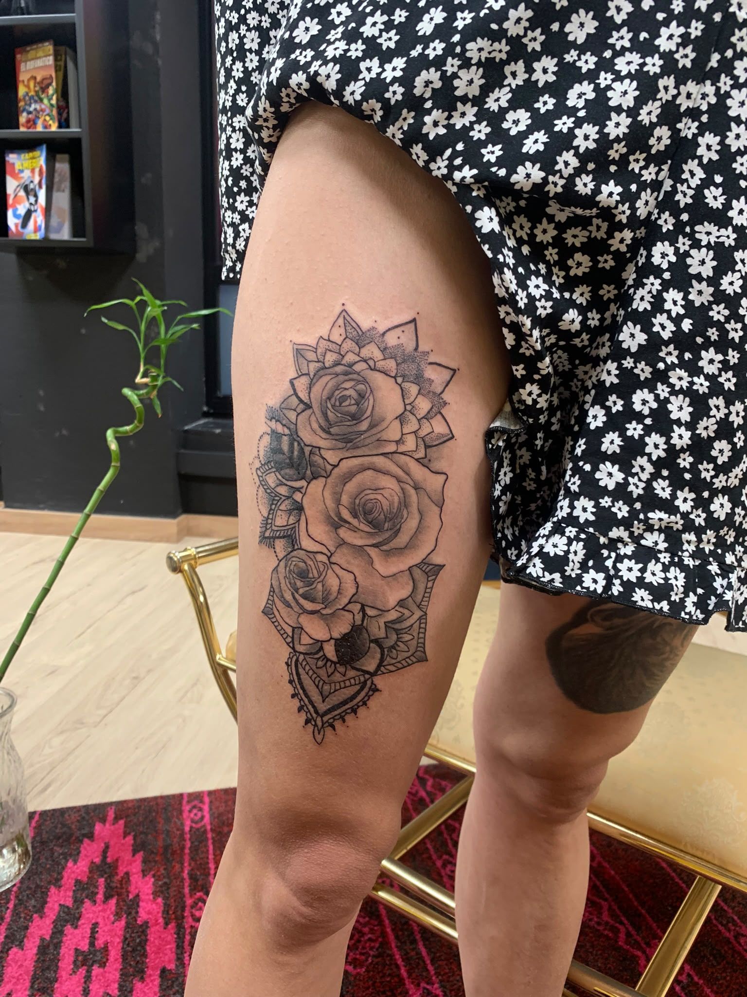 Primer plano de una pierna con un tatuaje floral negro, con un vestido negro estampado en un espacio interior.