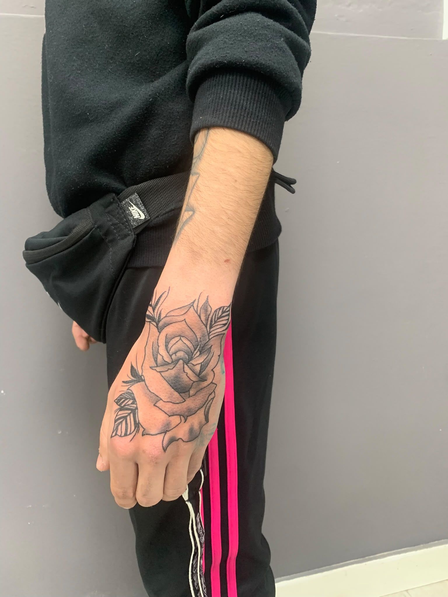 Brazo con un tatuaje de rosa junto a unos pantalones negros con franjas laterales de color rosa brillante, frente a una pared gris