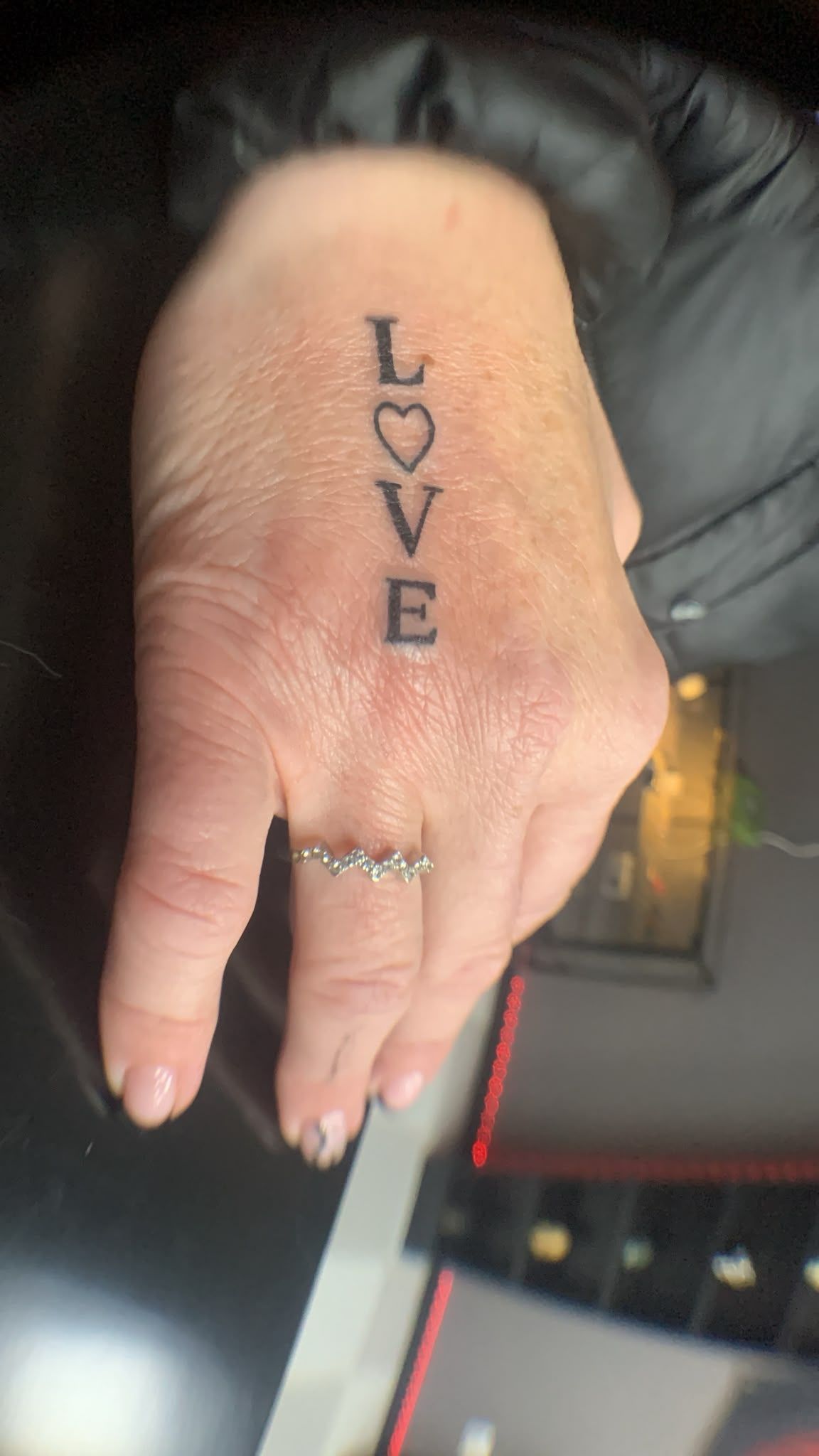 Una mano con «LOVE» tatuado en vertical en el dorso, con un anillo, apoyada sobre una superficie oscura.