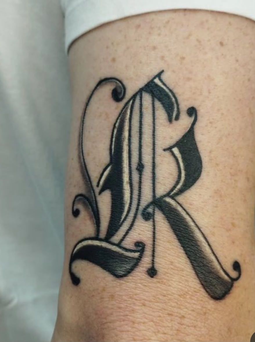 Tatuaje de la letra R ornamental en negro y gris sobre la piel, con adornos decorativos