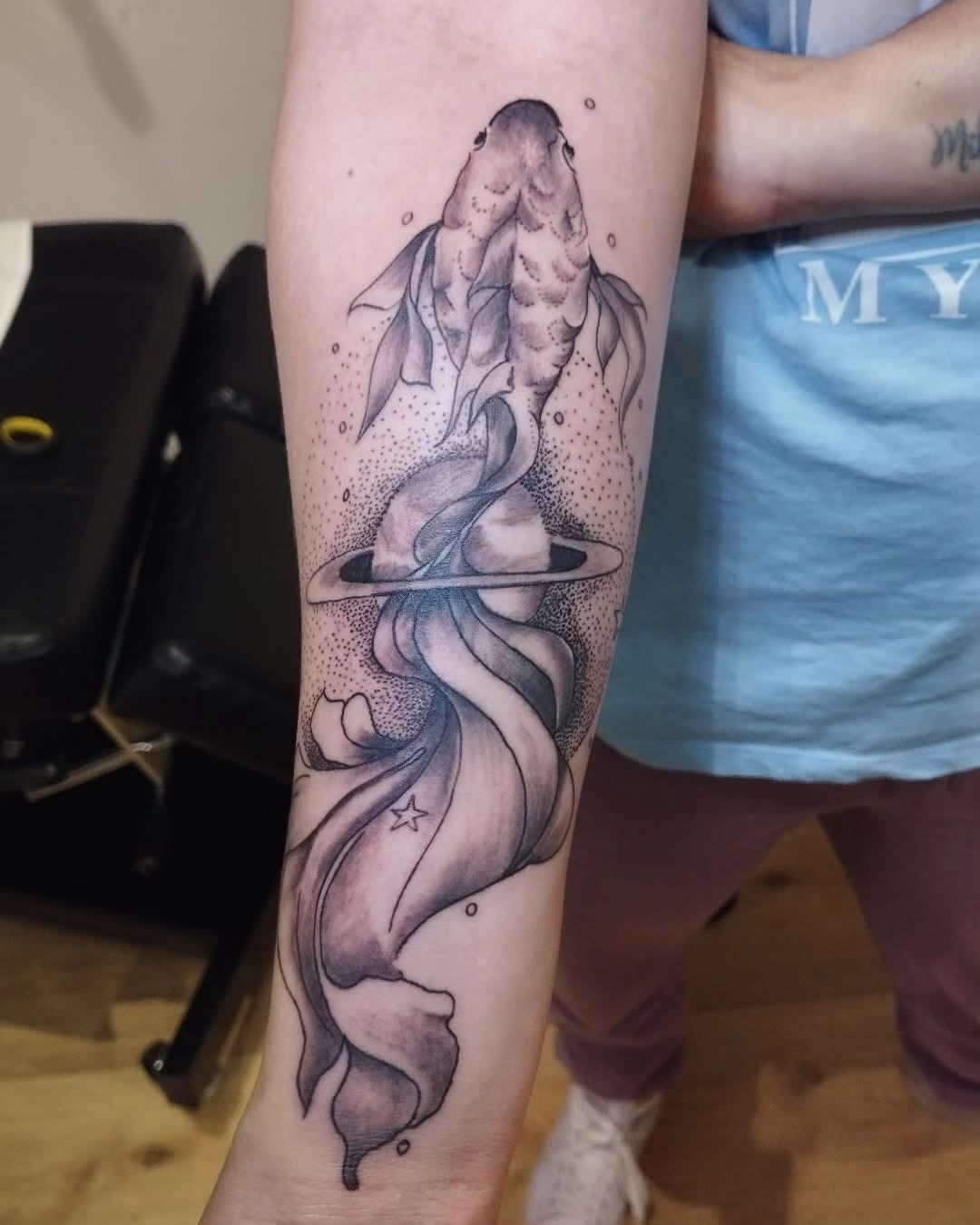 Tatuaje en negro y gris de una figura encapuchada en la parte superior del brazo, con formas fluidas parecidas al humo debajo.