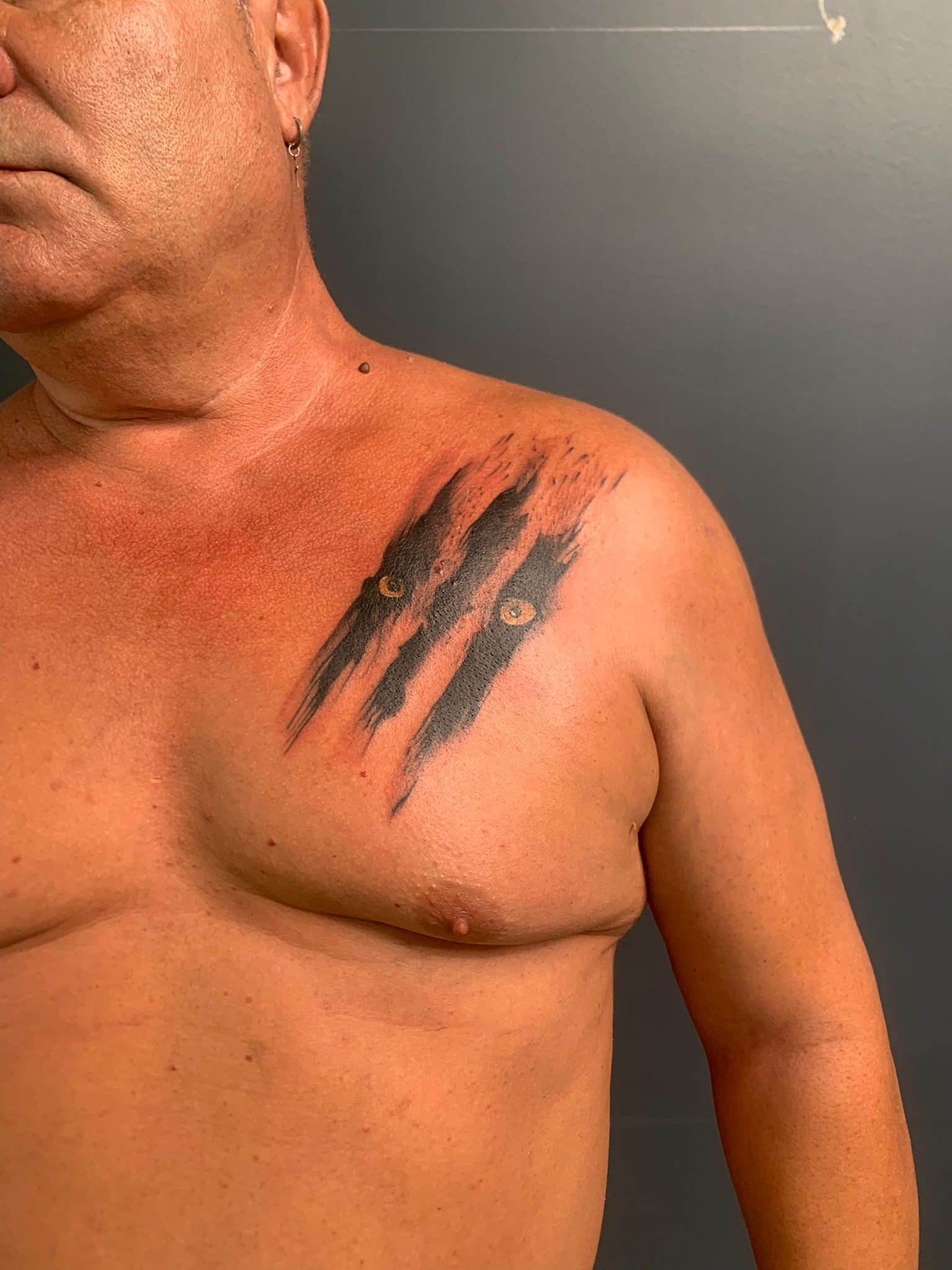 Torso desnudo de un hombre, con un gran tatuaje negro en forma de garra en el hombro izquierdo y el pecho, sobre un fondo gris