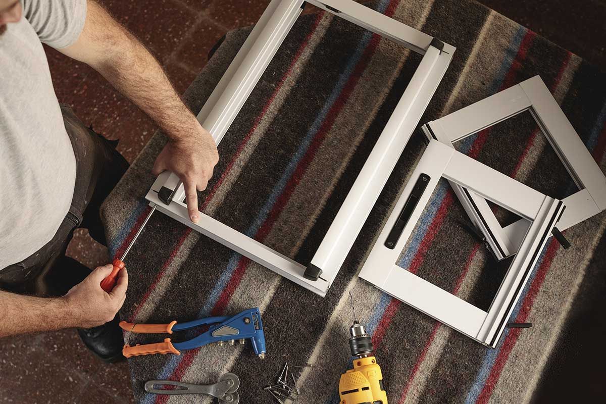 Persona ensamblando marco de ventana blanco sobre alfombra rayada con herramientas.