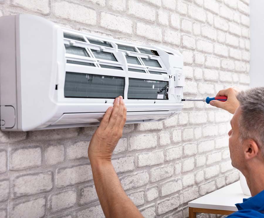 Una persona usa un destornillador para reparar un acondicionador de aire blanco montado en una pared de ladrillos.