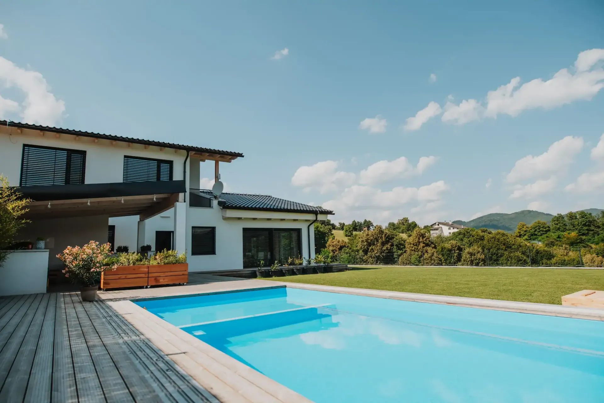Casa con piscina y terraza de madera en un día soleado. Césped verde y cielo azul con nubes.