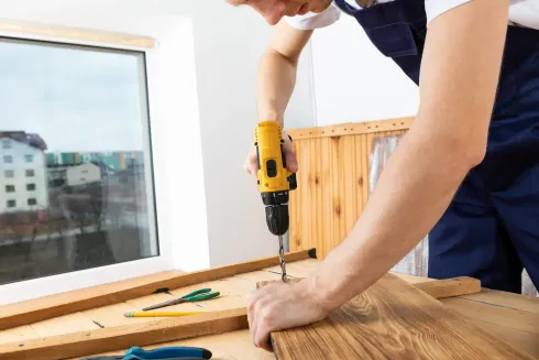 Persona que utiliza un taladro eléctrico amarillo sobre una superficie de madera cerca de una ventana.