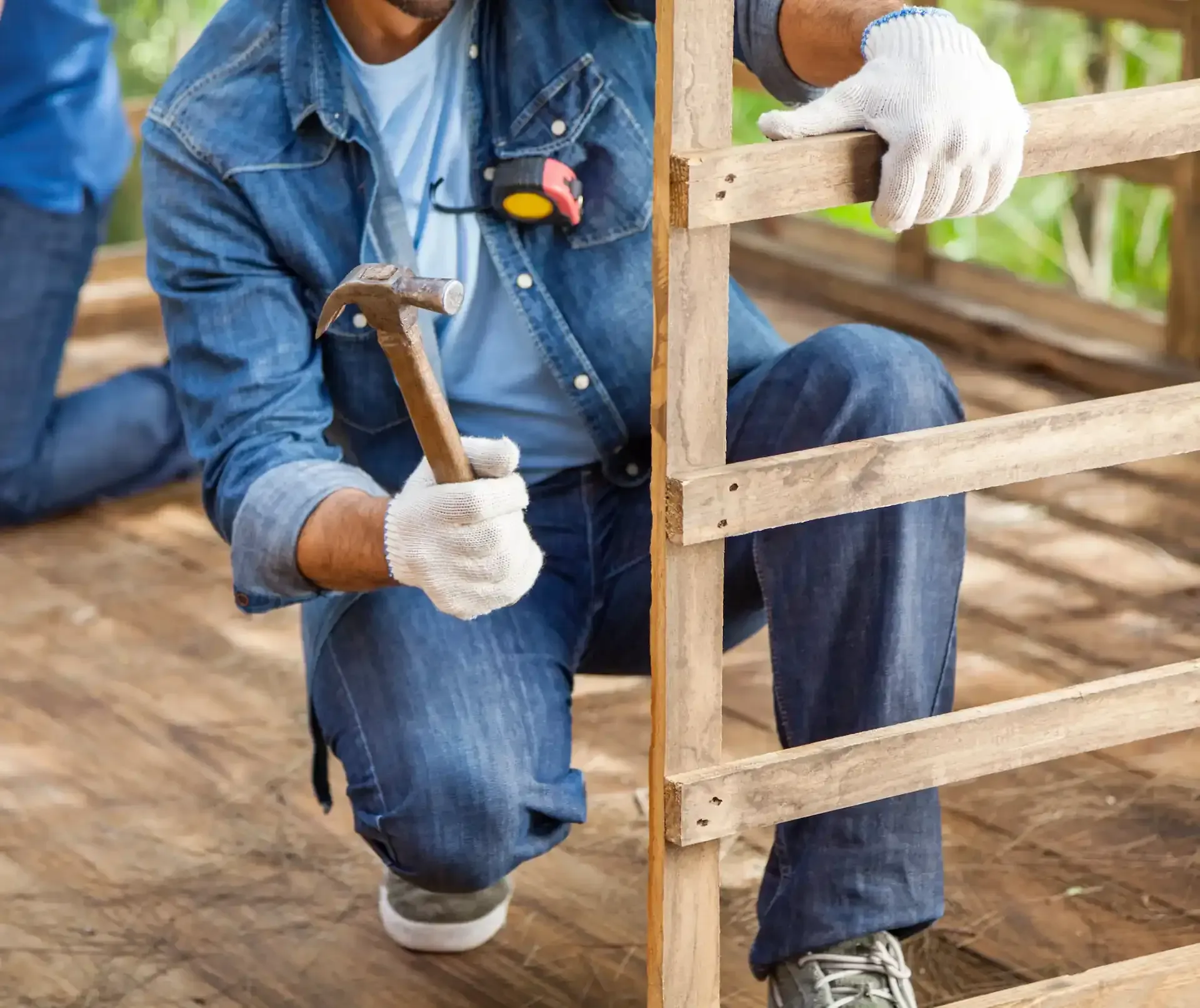 Persona arrodillada, martillando un tablón de madera, con guantes y ropa vaquera, sobre una plataforma de madera.