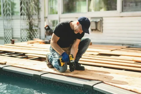 Hombre arrodillado, utilizando un taladro sobre tablones de madera cerca de una piscina.