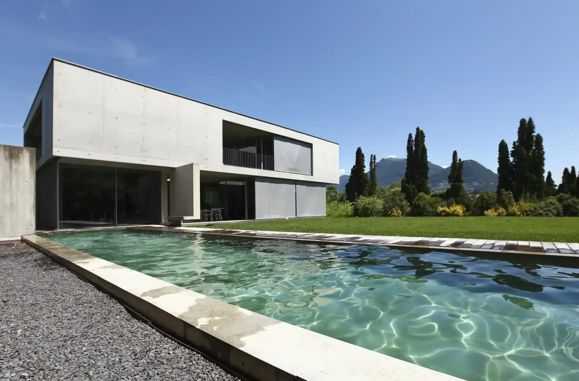Moderna casa de hormigón con piscina rectangular, rodeada de césped verde y árboles, bajo un cielo azul.