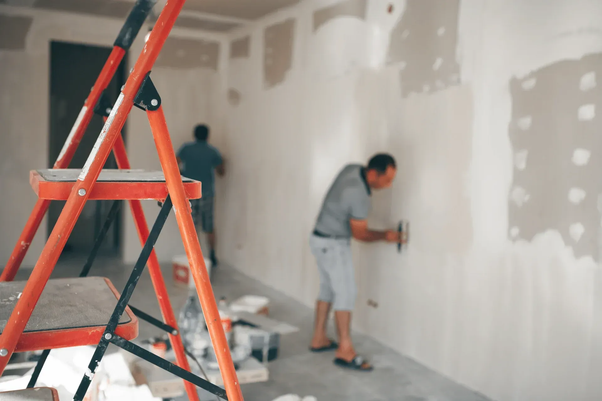 Obreros de la construcción enyesan una pared; en primer plano hay una escalera naranja.