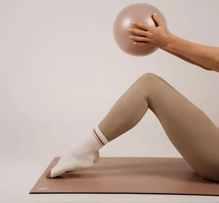 Una mujer está sentada en una esterilla de yoga sosteniendo una pelota rosa.
