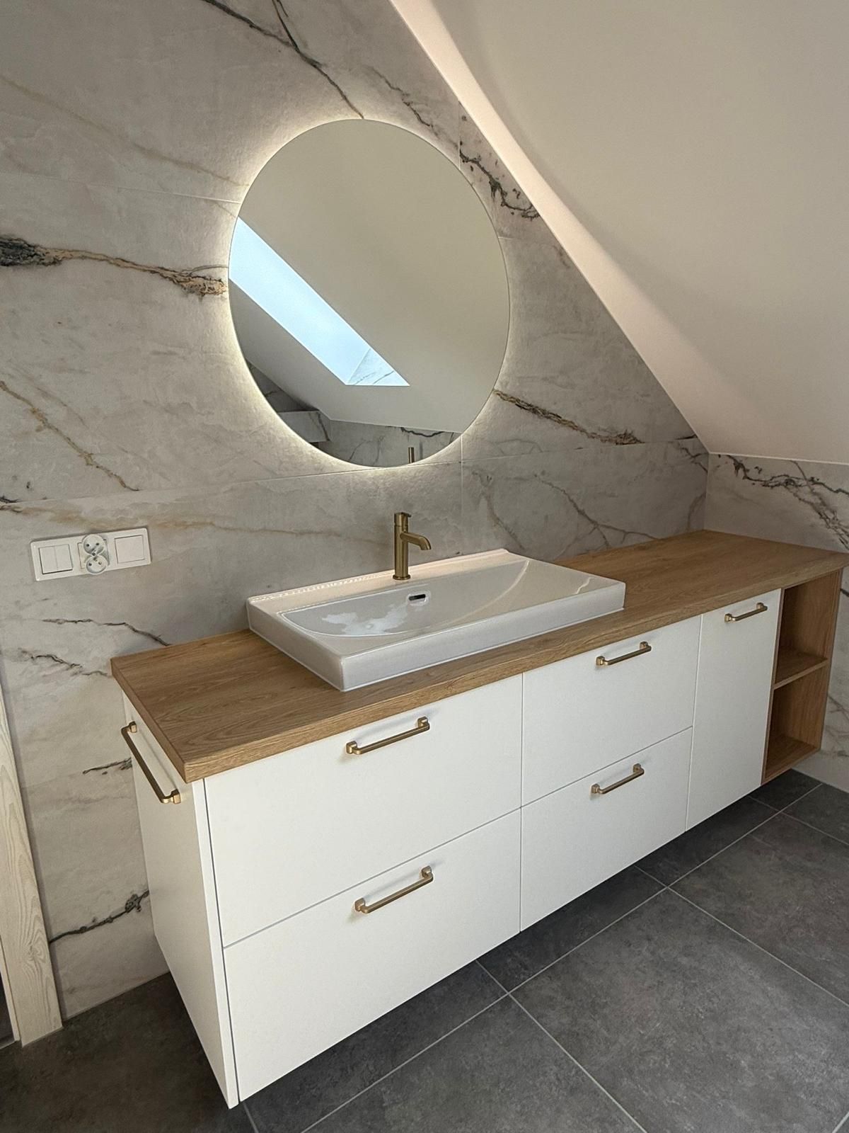 Moderner Badezimmerwaschtisch mit weißer Arbeitsplatte und Schubladen, rechteckigem Waschbecken, goldener Armatur und rundem, hinterleuchtetem Spiegel.