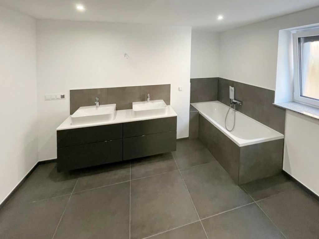 Ein modernes Badezimmer mit einem dunklen Doppelwaschtisch, einer gefliesten Badewanne vor einer grauen Akzentwand und grauen Bodenfliesen.