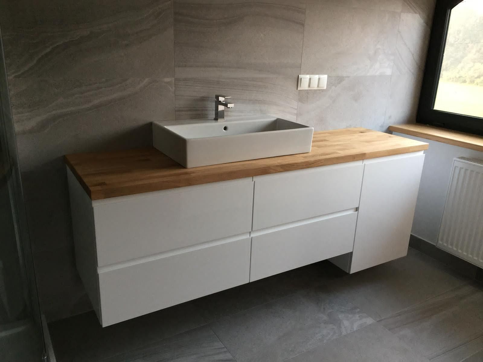 Ein moderner Badezimmerwaschtisch mit weißem rechteckigem Waschbecken auf einer Holzarbeitsplatte und weißen Schubladen vor einer grau gefliesten Wand.