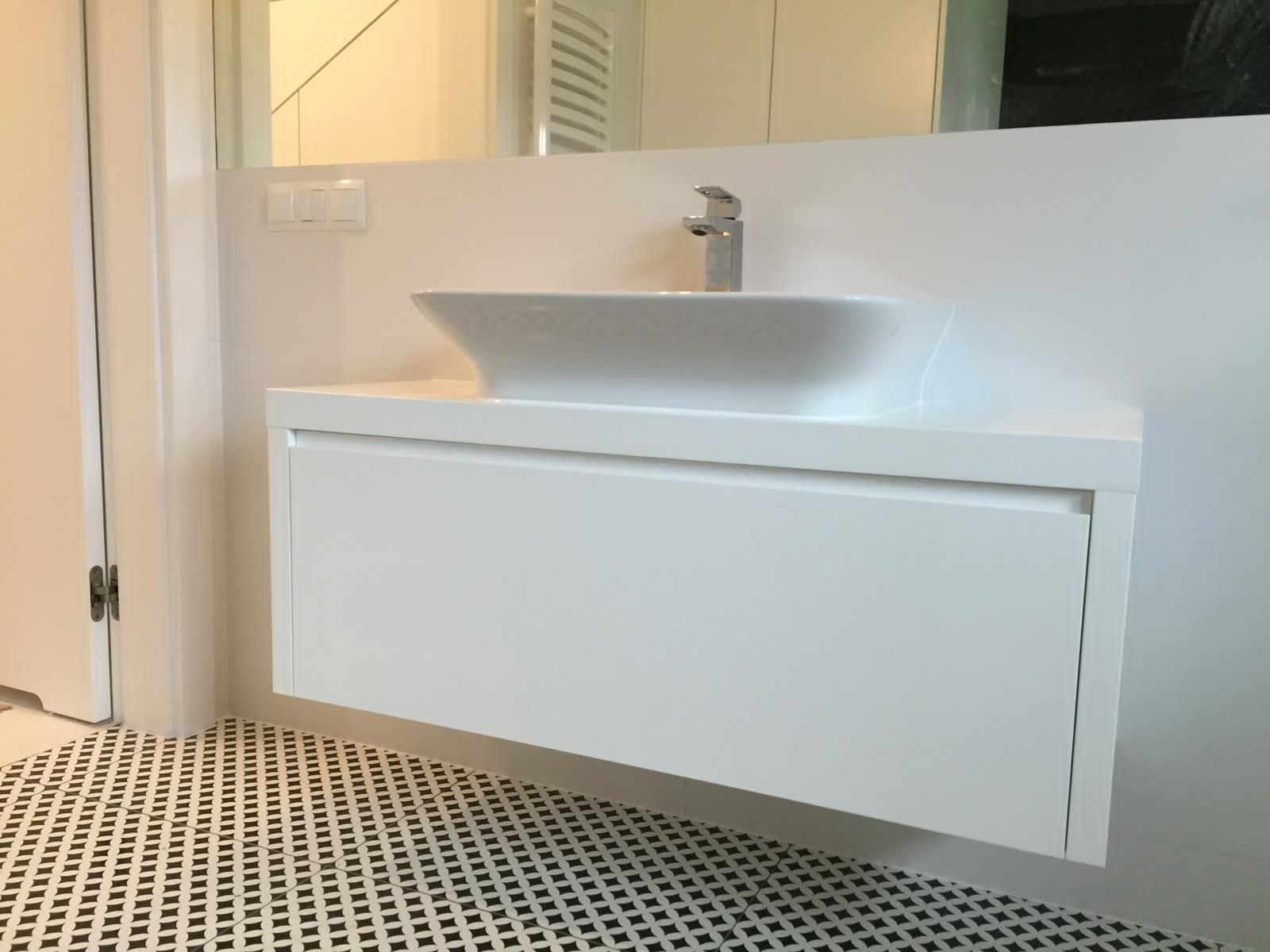 Ein weißer, schwebender Waschtisch mit quadratischem Aufsatzwaschbecken und Chromarmatur vor einer schlichten Wand über gemusterten Fliesen.