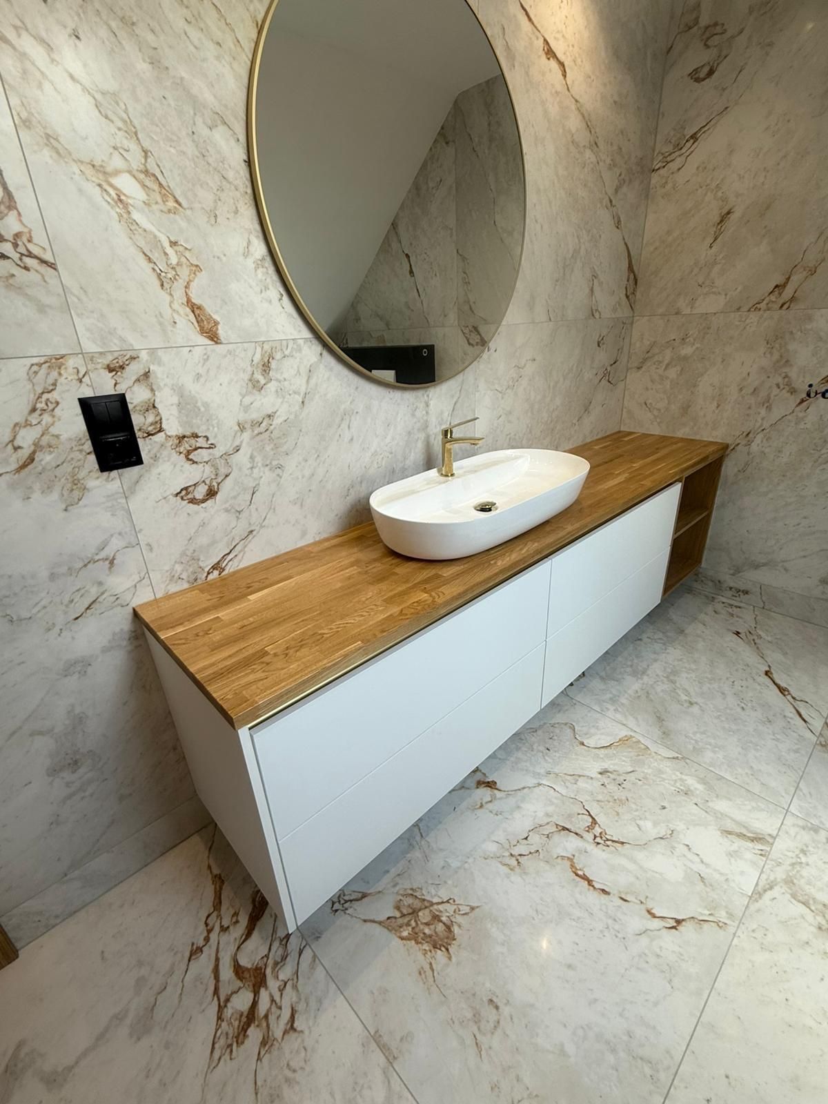 Ein weißer Badezimmerwaschtisch mit Holzplatte und ovalem Waschbecken, der an einer Wand mit weißen und goldenen Marmorfliesen steht.