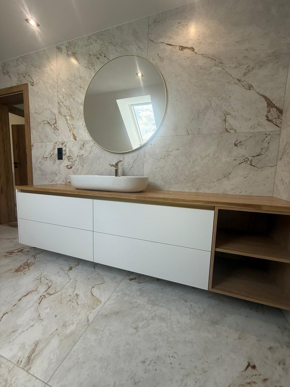 Ein schwebender Waschtisch mit weißem Waschbecken und rundem Spiegel vor einer Wand aus Marmorfliesen.