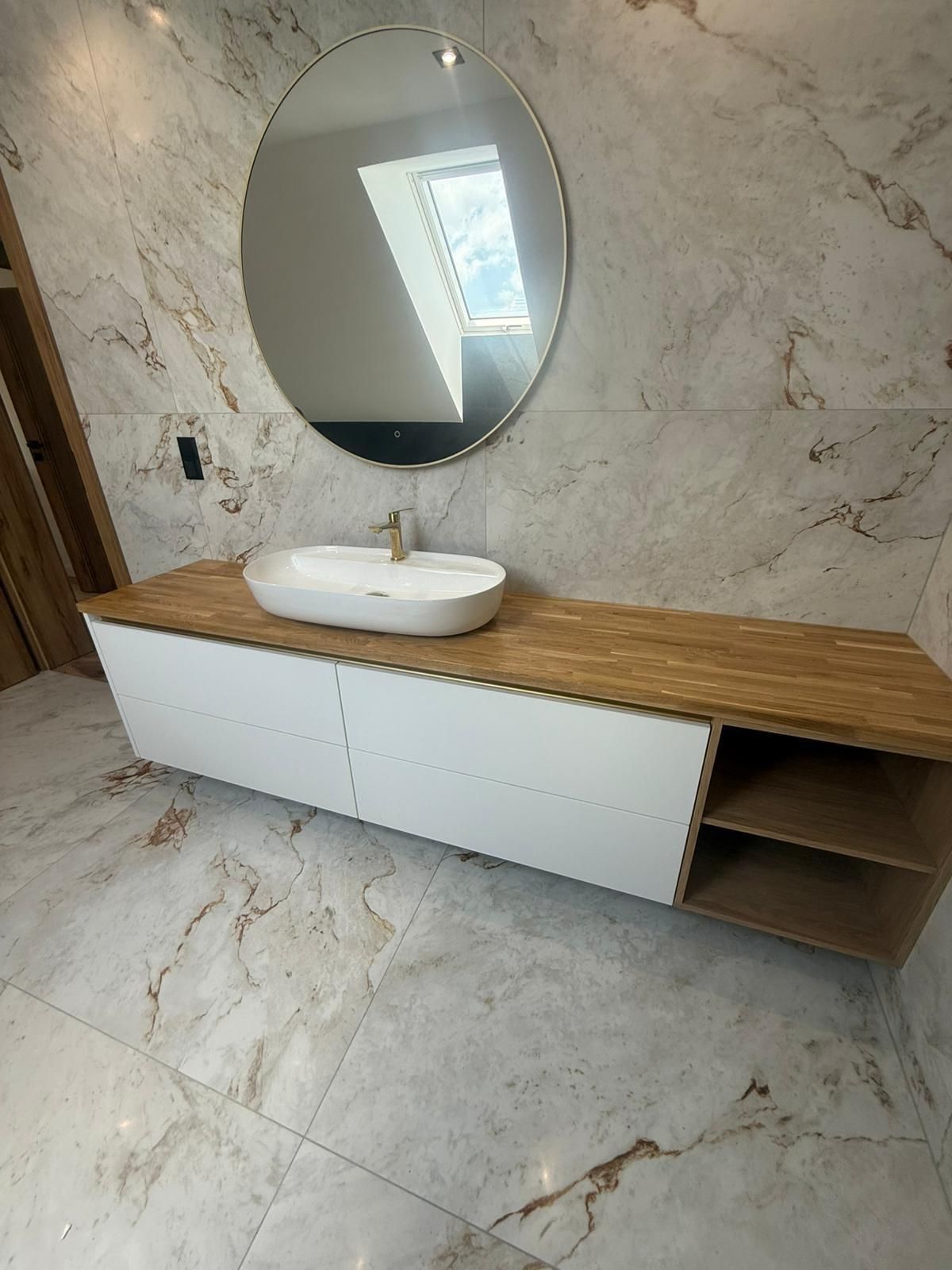 Ein Badezimmerschrank mit weißem Aufsatzwaschbecken, hölzerner Arbeitsplatte, weißen Schränken und einem ovalen Spiegel an einer marmorgefliesten Wand.