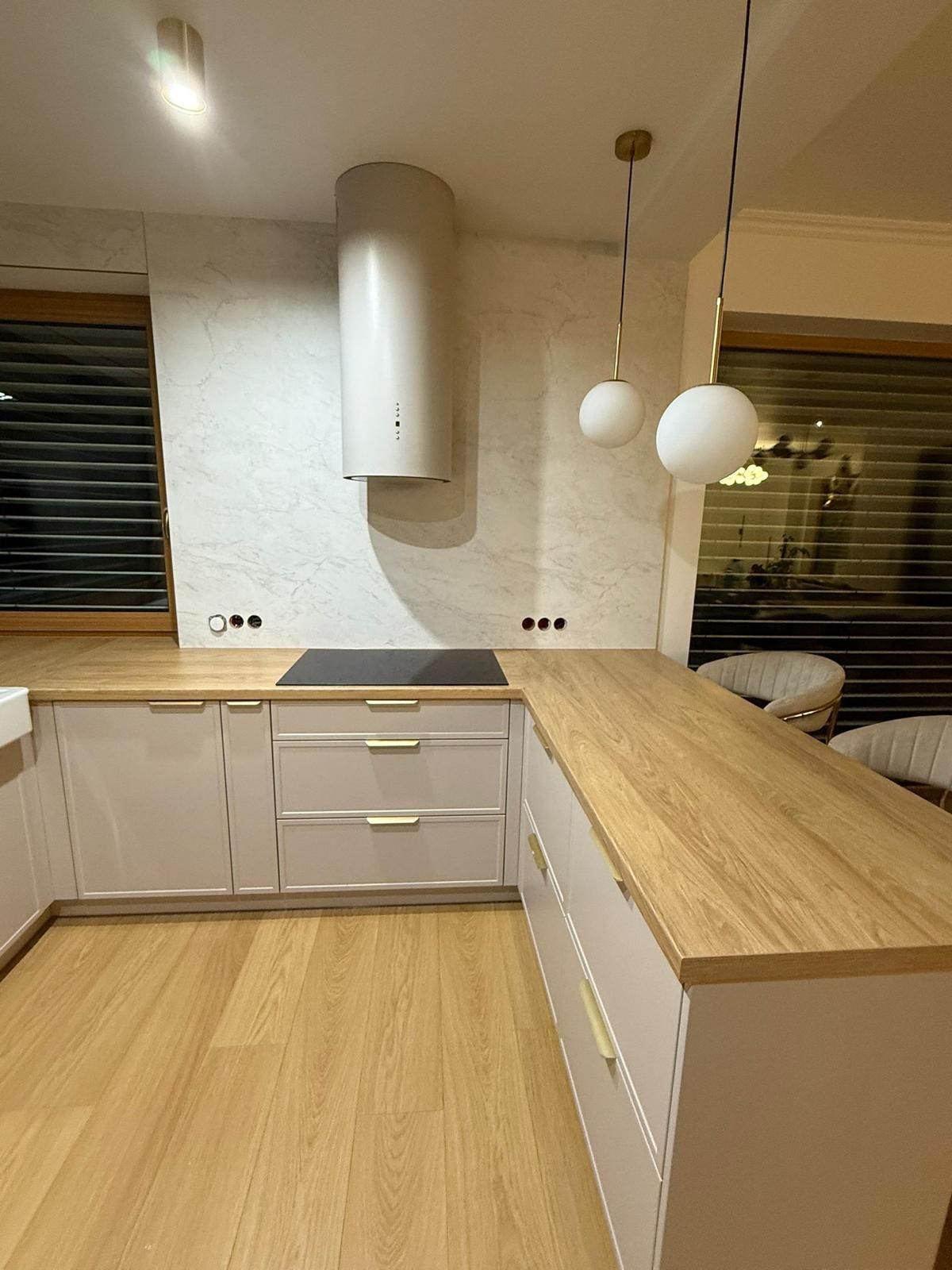 Moderne Küche mit weißen Schränken, holzfarbenen Arbeitsplatten, einer zylindrischen Dunstabzugshaube und zwei hängenden Kugelpendelleuchten.