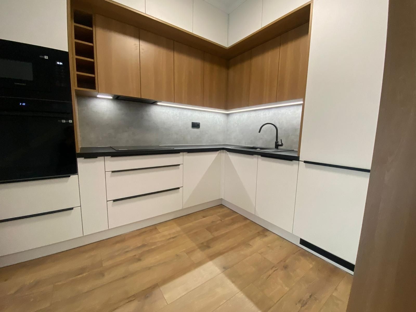 Moderne L-förmige Küche mit weißen Unterschränken, holzfarbenen Oberschränken, schwarzer Arbeitsplatte und grauem Fliesenspiegel.