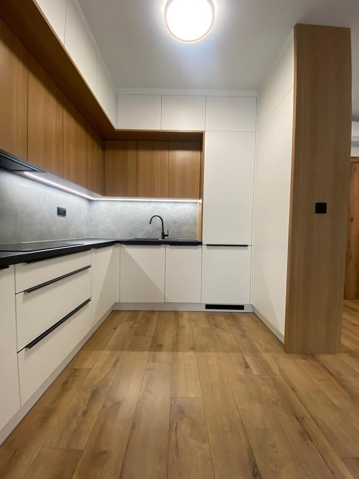 Moderne Küche mit weißen Unterschränken, holzfarbenen Oberschränken, schwarzen Beschlägen und hellem Holzboden.