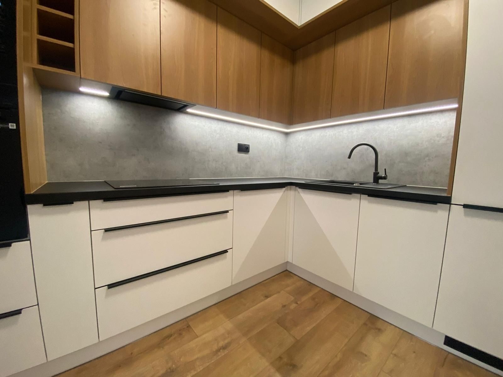 Eine moderne L-förmige Küche mit weißen Unterschränken, hellen Holzoberschränken, einer dunklen Arbeitsplatte und Unterbaubeleuchtung.