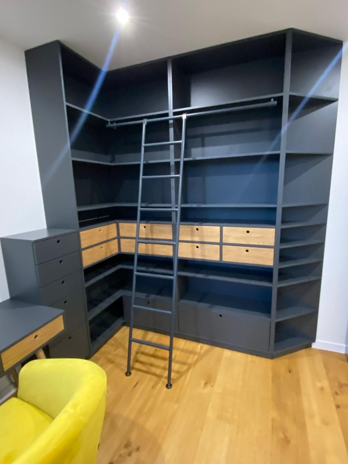 Ein dunkelgraues Eckregal mit Holzschubladen und integrierter Rollleiter, daneben ein passender Schreibtisch und Stuhl.