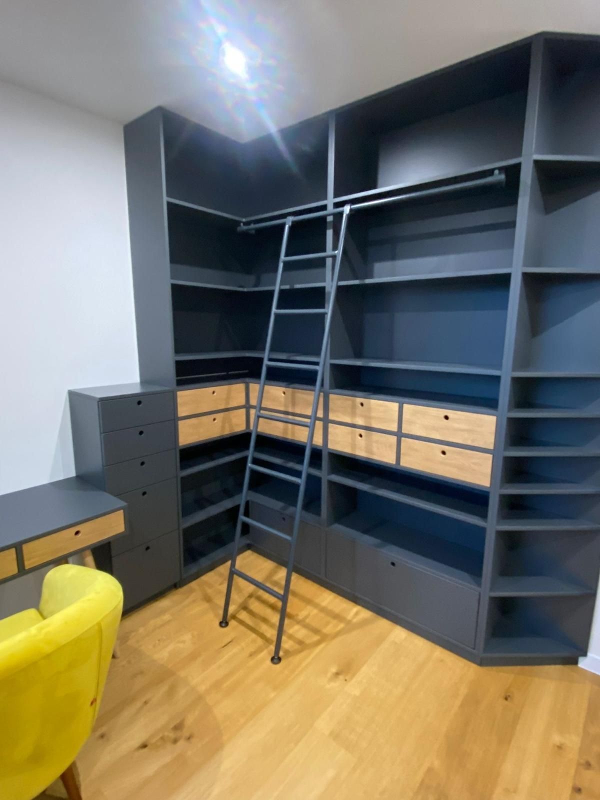 Eine dunkelgraue, L-förmige Bibliothekswand mit eingebauten Holzschubladen, einer ausziehbaren Metallleiter und einem passenden Schreibtisch.