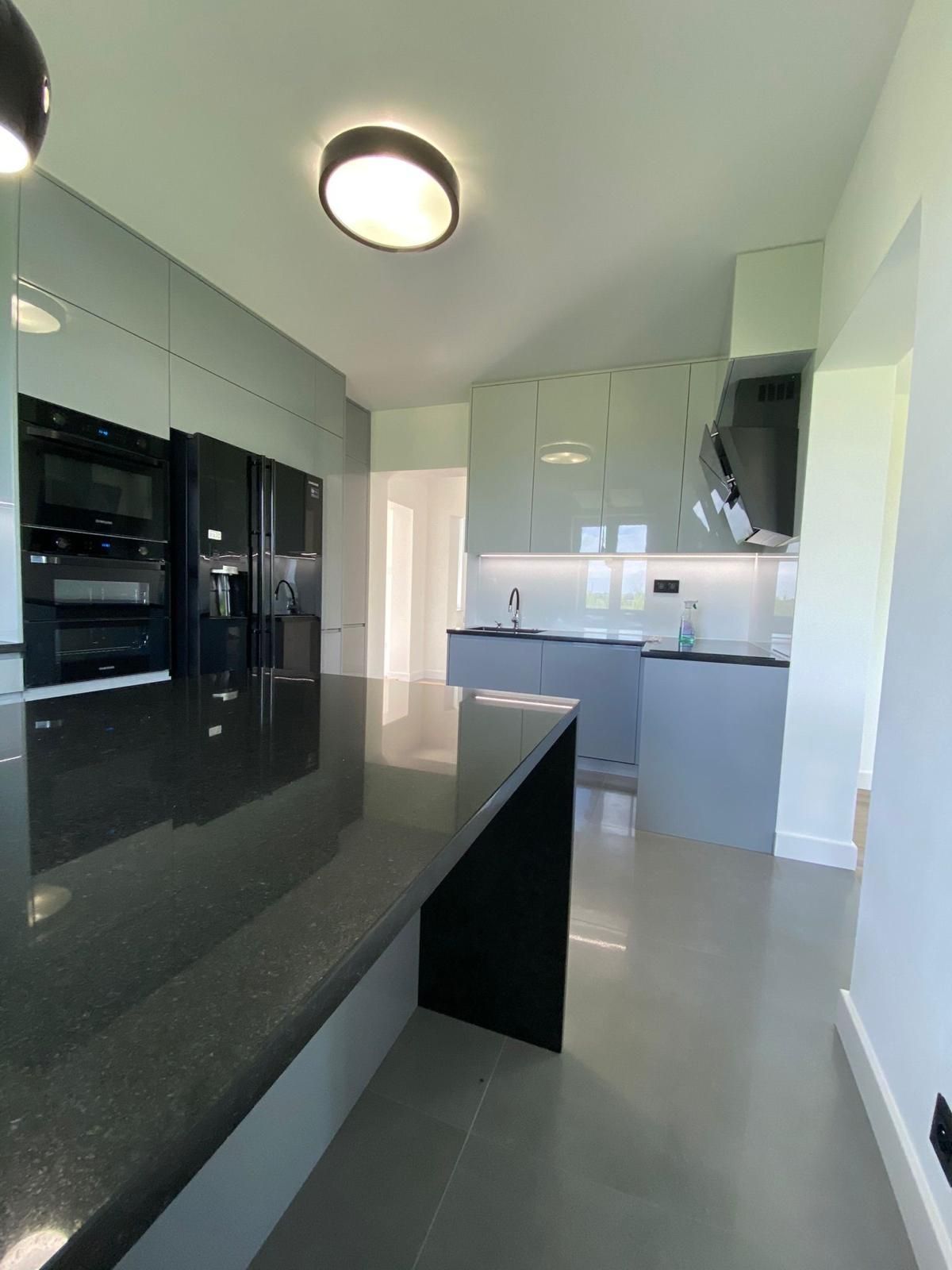 Moderne Küche mit schwarzer Granitinsel, eingebauten Edelstahlöfen, grauen Schränken und glänzenden hellgrauen Böden.