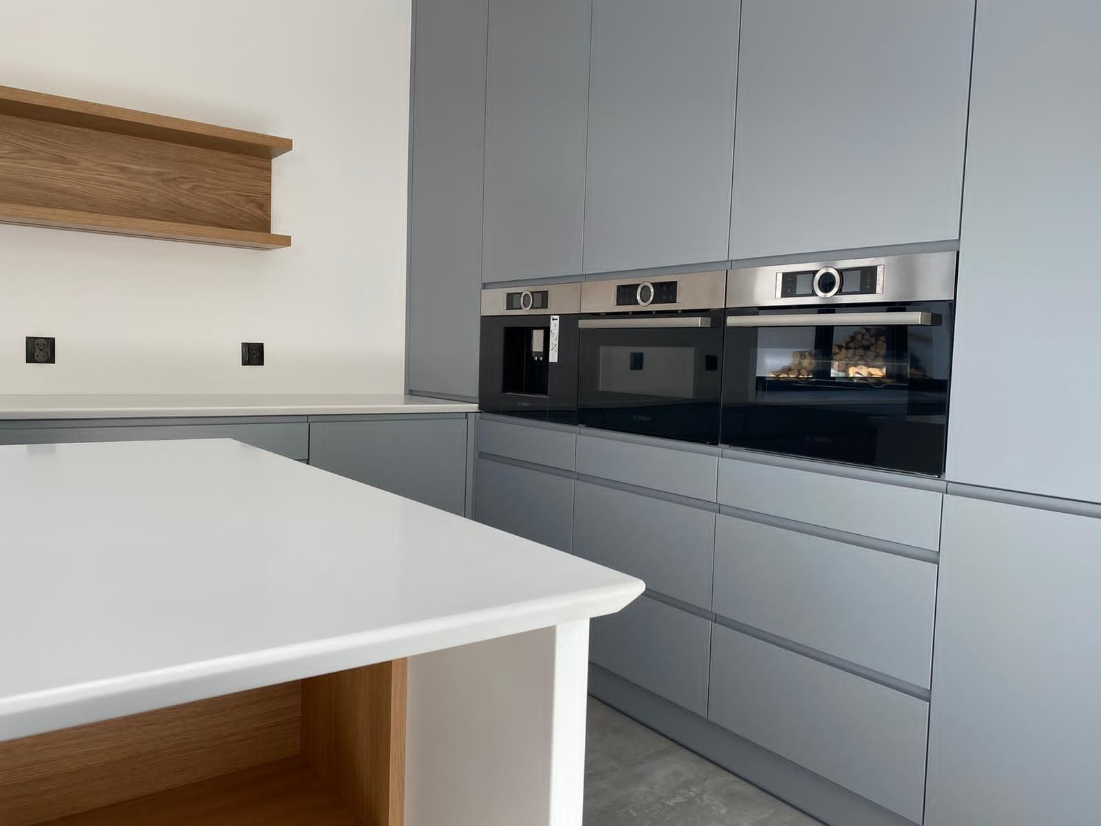 Eine moderne Küche mit grauen Schränken, einer weißen Kochinsel, Holzregalen und eingebauten Edelstahlgeräten.