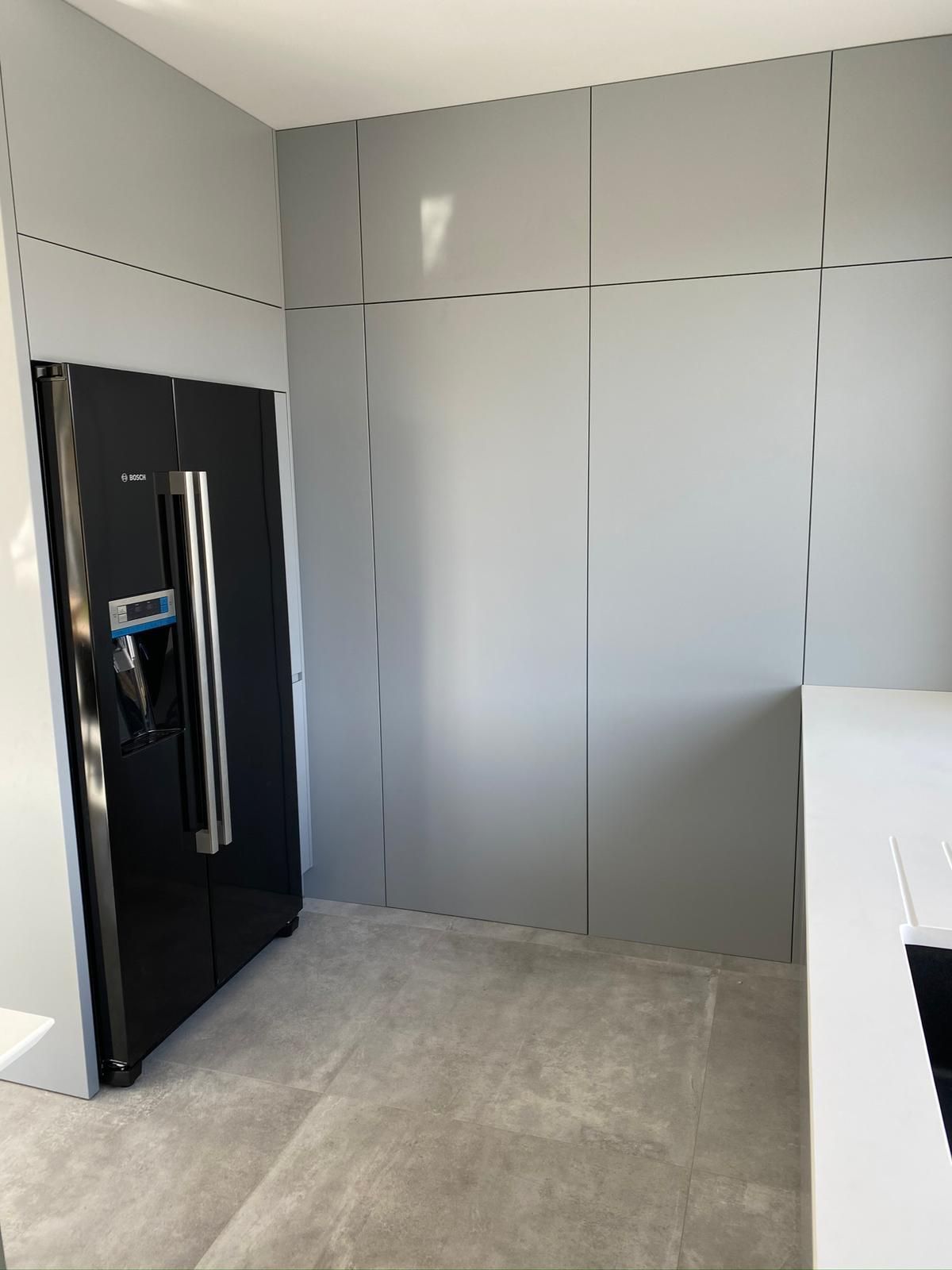 Ein schwarzer Side-by-Side-Kühlschrank neben einer Wand aus hohen, grauen Schränken mit flachen Fronten in einer modernen, hellgrau gefliesten Küche.