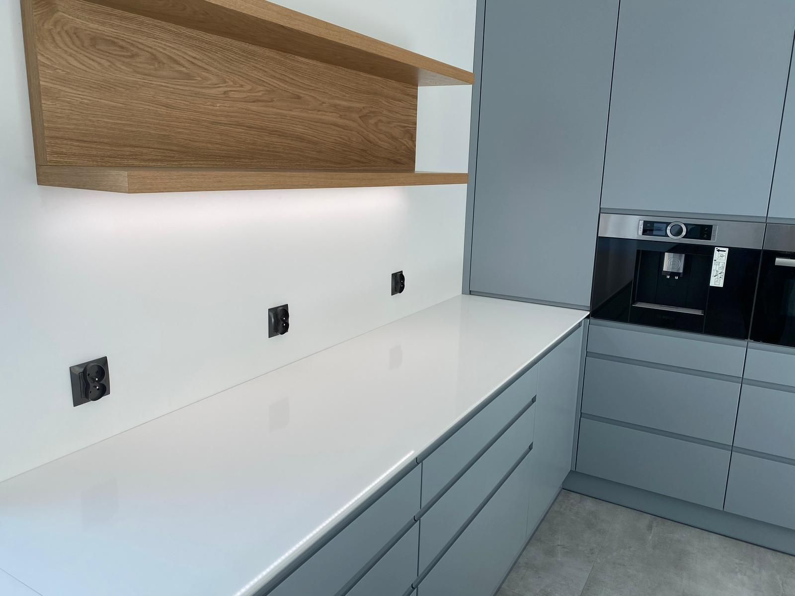 Eine moderne Küche mit hellgrauen Schränken, einer weißen Arbeitsplatte, schwebenden Holzregalen und einer integrierten Kaffeemaschine.