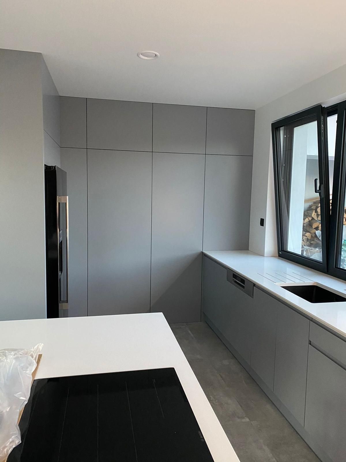 Eine moderne Küche mit mattgrauen Schränken, einer weißen Kochinsel, weißen Arbeitsplatten und einem schwarzen Fensterrahmen.