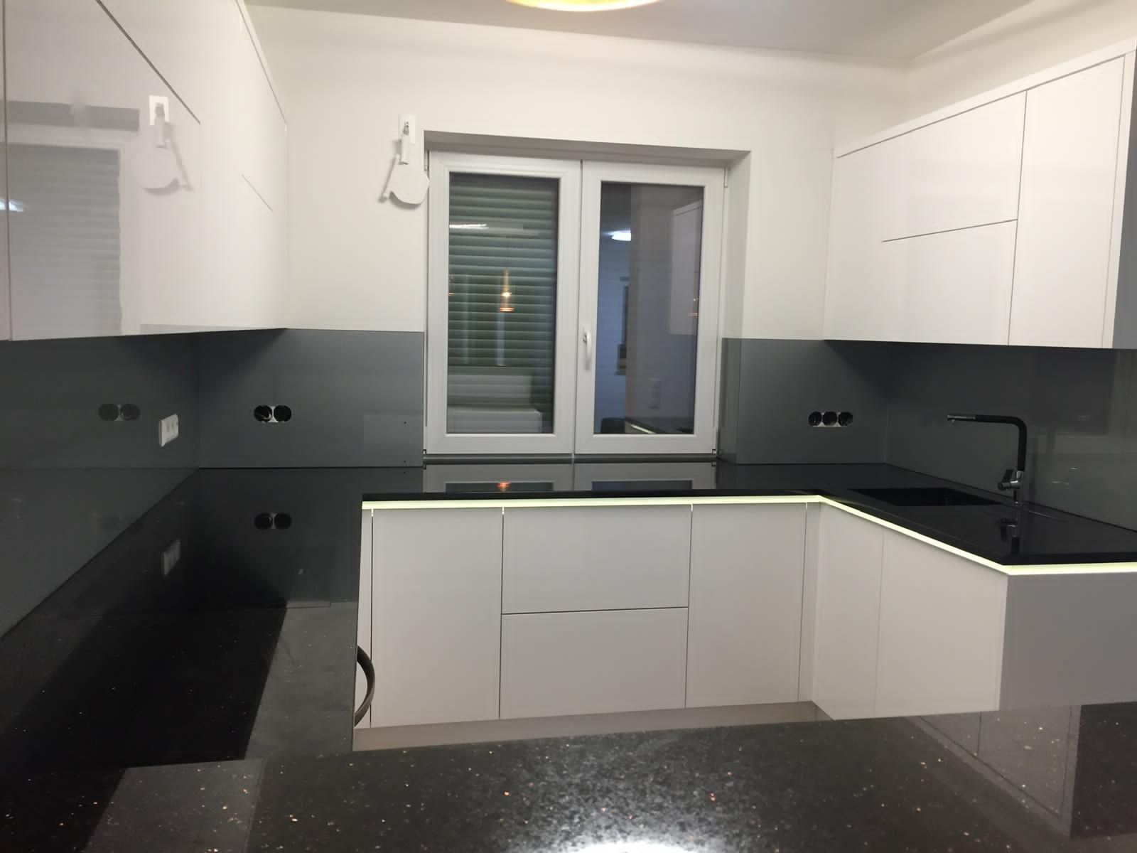 Moderne L-förmige Küche mit weißen Schränken, glänzend schwarzen Arbeitsplatten und grauem Fliesenspiegel sowie einem zentralen Fenster.