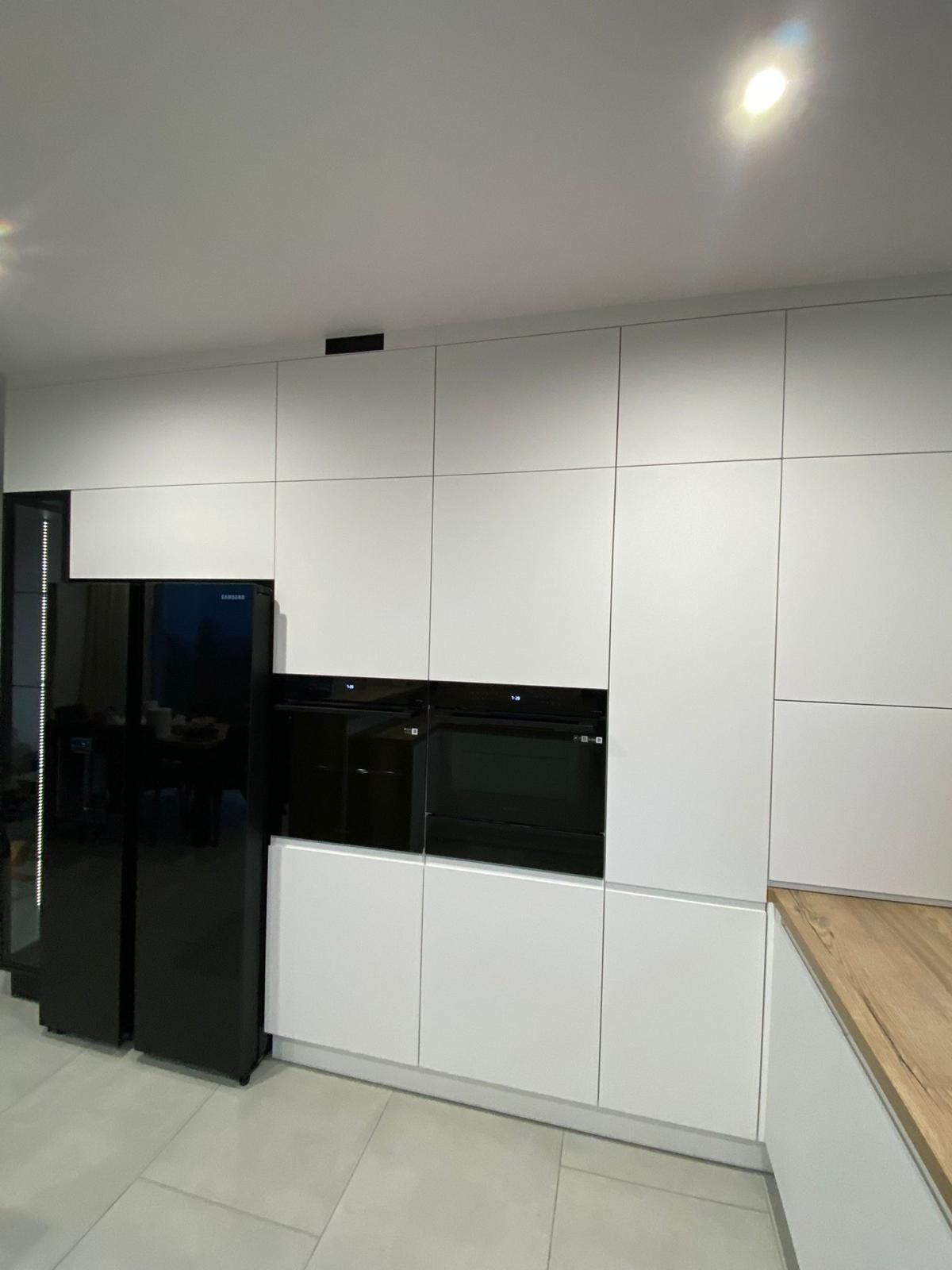 Eine minimalistische Küchenwand mit weißen, grifflosen Schränken, einem schwarzen Kühlschrank und eingebauten schwarzen Backöfen.