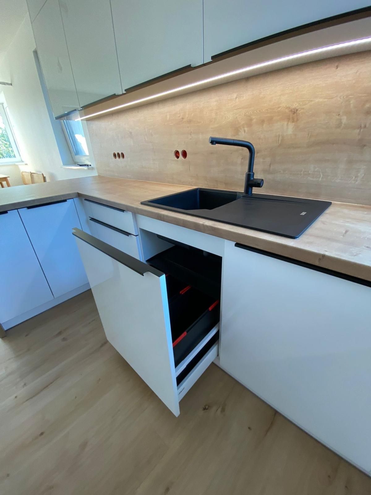 Moderne Küche mit weißen Schränken, hellen Holzarbeitsplatten, schwarzem Spülbecken und Wasserhahn sowie einer teilweise offenen Abfallschublade.